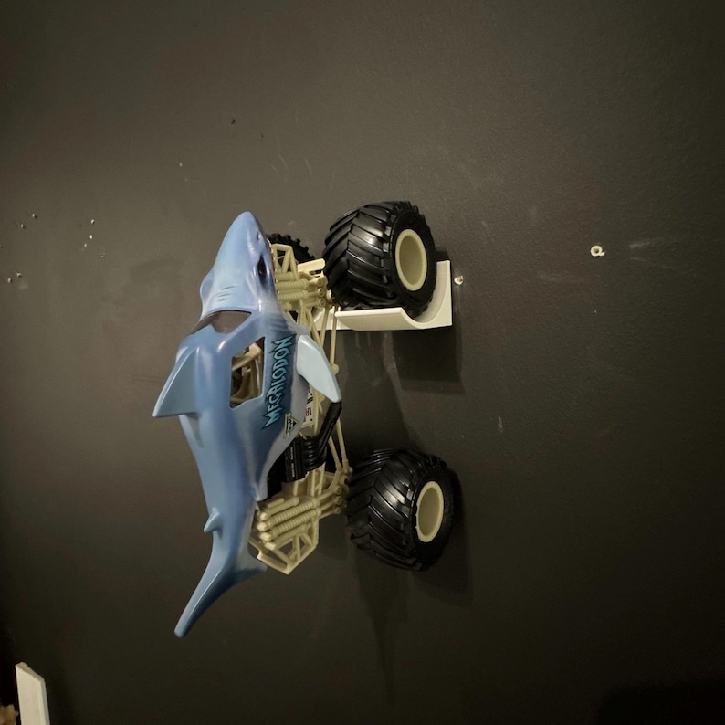 May include: A blue and white Megalodon monster truck toy with large black tires. The truck is mounted on a white shelf against a dark gray wall. The text "MEGALODON" is visible on the side of the truck.