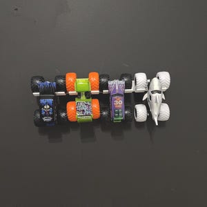 Puede incluir: Una colecci&oacute;n de cinco camiones monstruo de juguete. Los camiones tienen neum&aacute;ticos grandes y dise&ntilde;os coloridos. Un cami&oacute;n es azul con un gr&aacute;fico de calavera, otro es verde con neum&aacute;ticos naranjas y un tercero es morado con el n&uacute;mero 30.