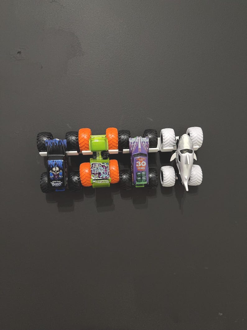 Puede incluir: Una colecci&oacute;n de cinco camiones monstruo de juguete. Los camiones tienen neum&aacute;ticos grandes y dise&ntilde;os coloridos. Un cami&oacute;n es azul con un gr&aacute;fico de calavera, otro es verde con neum&aacute;ticos naranjas y un tercero es morado con el n&uacute;mero 30.