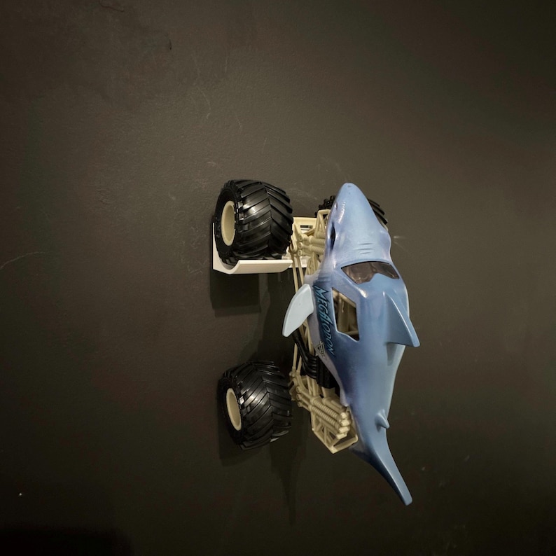 May include: A blue and white toy monster truck shaped like a shark. The truck has large black tires and the word "Megalodon" printed on the side. The toy is set against a dark background.