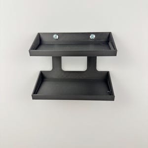 May include: A black, two-tiered wall-mounted shelf. The shelf has two rectangular trays, one above the other, connected by a central support. Two screws secure the top tray to the wall. The shelf is made of a matte black material.