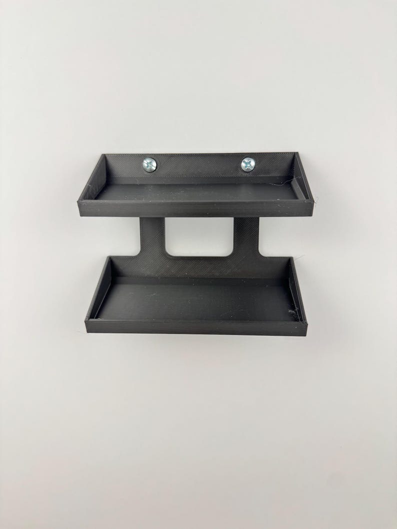 May include: A black, two-tiered wall-mounted shelf. The shelf has two rectangular trays, one above the other, connected by a central support. Two screws secure the top tray to the wall. The shelf is made of a matte black material.