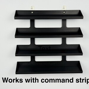 May include: A black, four-tiered wall shelf with a minimalist design. The shelves are rectangular with raised edges. The text "Text" is visible on the third shelf, and the text "Works with command strips" is at the bottom.