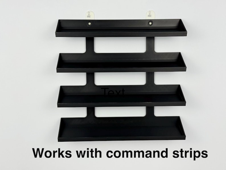 May include: A black, four-tiered wall shelf with a minimalist design. The shelves are rectangular with raised edges. The text "Text" is visible on the third shelf, and the text "Works with command strips" is at the bottom.