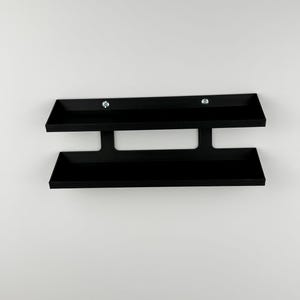 May include: A black, wall-mounted shelf with two horizontal trays connected by a vertical support. The shelf is made of a matte black material and is mounted on a white wall. Two silver screws secure the top tray.