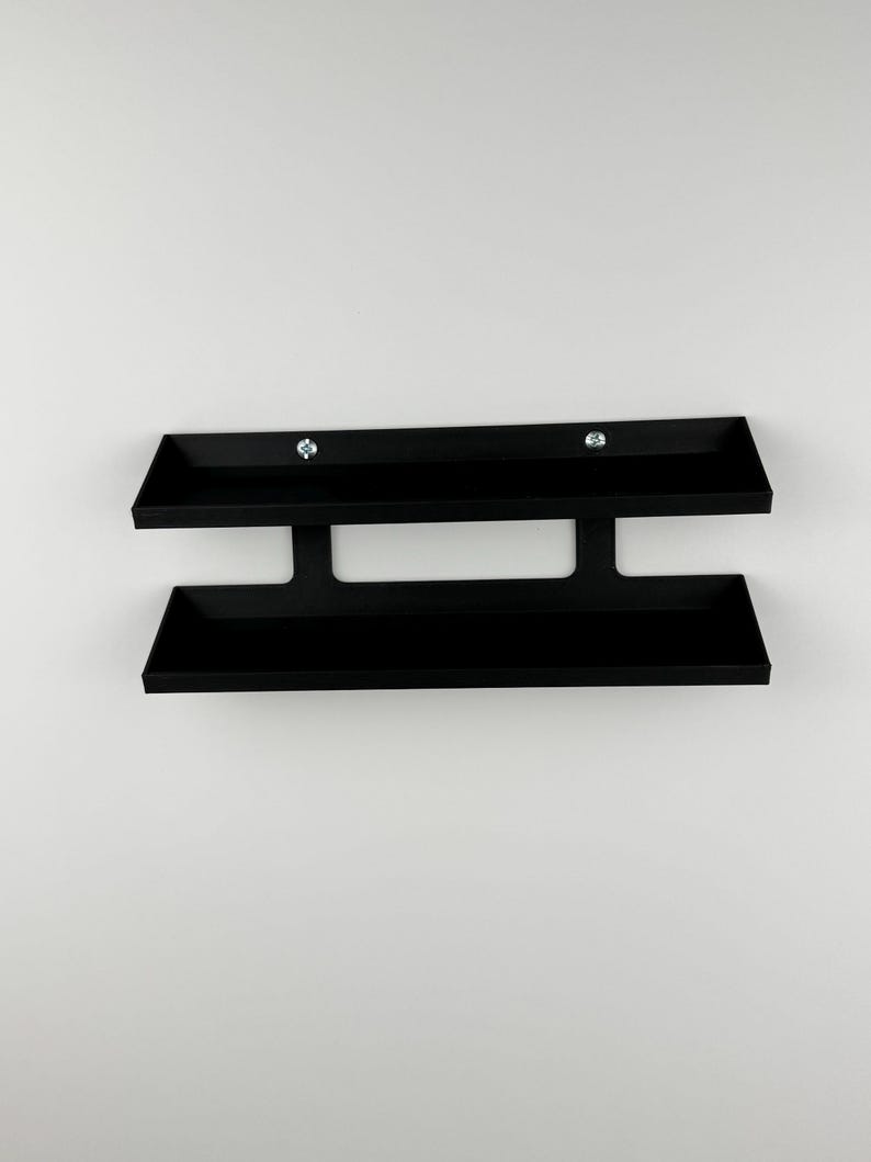 May include: A black, wall-mounted shelf with two horizontal trays connected by a vertical support. The shelf is made of a matte black material and is mounted on a white wall. Two silver screws secure the top tray.