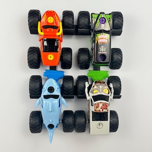May include: Four toy monster trucks in a row. The trucks are red, green, blue, and white. The red truck has a flame design. The green truck has the words "Grave Digger" on it. The blue truck is shaped like a shark.