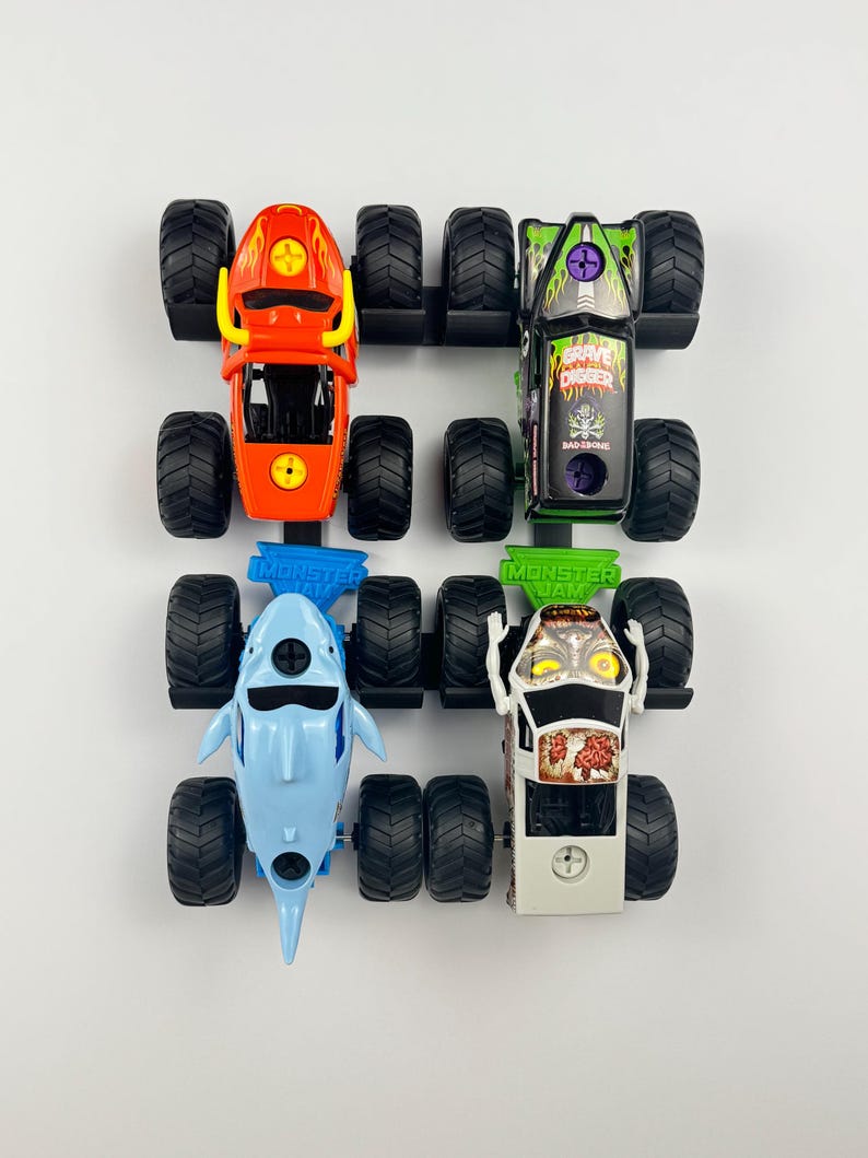 May include: Four toy monster trucks in a row. The trucks are red, green, blue, and white. The red truck has a flame design. The green truck has the words "Grave Digger" on it. The blue truck is shaped like a shark.