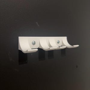 Puede incluir: Un perchero blanco montado en la pared con cuatro ganchos curvos. El perchero est&aacute; hecho de pl&aacute;stico y est&aacute; fijado a una pared oscura con tornillos. Los ganchos est&aacute;n espaciados uniformemente y dise&ntilde;ados para colgar objetos.