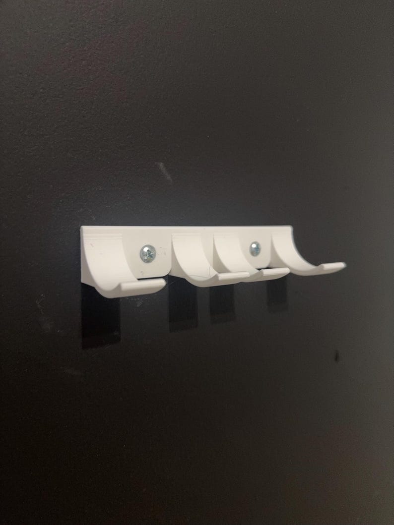 Puede incluir: Un perchero blanco montado en la pared con cuatro ganchos curvos. El perchero est&aacute; hecho de pl&aacute;stico y est&aacute; fijado a una pared oscura con tornillos. Los ganchos est&aacute;n espaciados uniformemente y dise&ntilde;ados para colgar objetos.