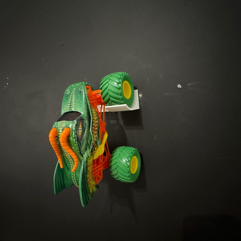 May include: Green and orange monster truck toy with large green tires and yellow rims. The truck features a detailed design with orange tentacles and a textured body. The toy is mounted on a white shelf against a dark gray wall.