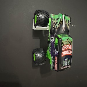May include: A toy monster truck in green and black, featuring the words "Grave Digger" and a skull graphic. The truck has large black tires with green accents and a flame design.