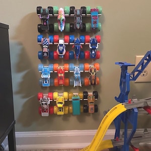 Puede incluir: Una colecci&oacute;n de camiones monstruo de juguete coloridos exhibidos en un estante montado en la pared. Los camiones presentan varios dise&ntilde;os y colores, incluyendo rojo, azul, verde y amarillo. Tambi&eacute;n se muestra una rampa de juguete amarilla y azul.