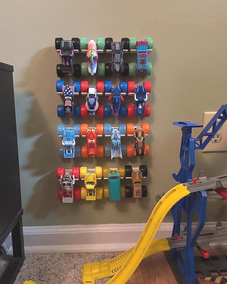Puede incluir: Una colecci&oacute;n de camiones monstruo de juguete coloridos exhibidos en un estante montado en la pared. Los camiones presentan varios dise&ntilde;os y colores, incluyendo rojo, azul, verde y amarillo. Tambi&eacute;n se muestra una rampa de juguete amarilla y azul.