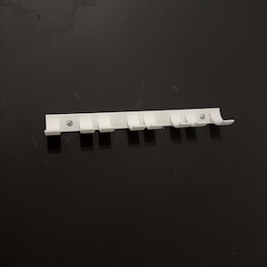 Puede incluir: Un organizador de pared de pl&aacute;stico blanco con siete ganchos. El organizador es rectangular y tiene una serie de ganchos peque&ntilde;os y espaciados para colgar objetos. Dos tornillos lo aseguran a la pared negra.
