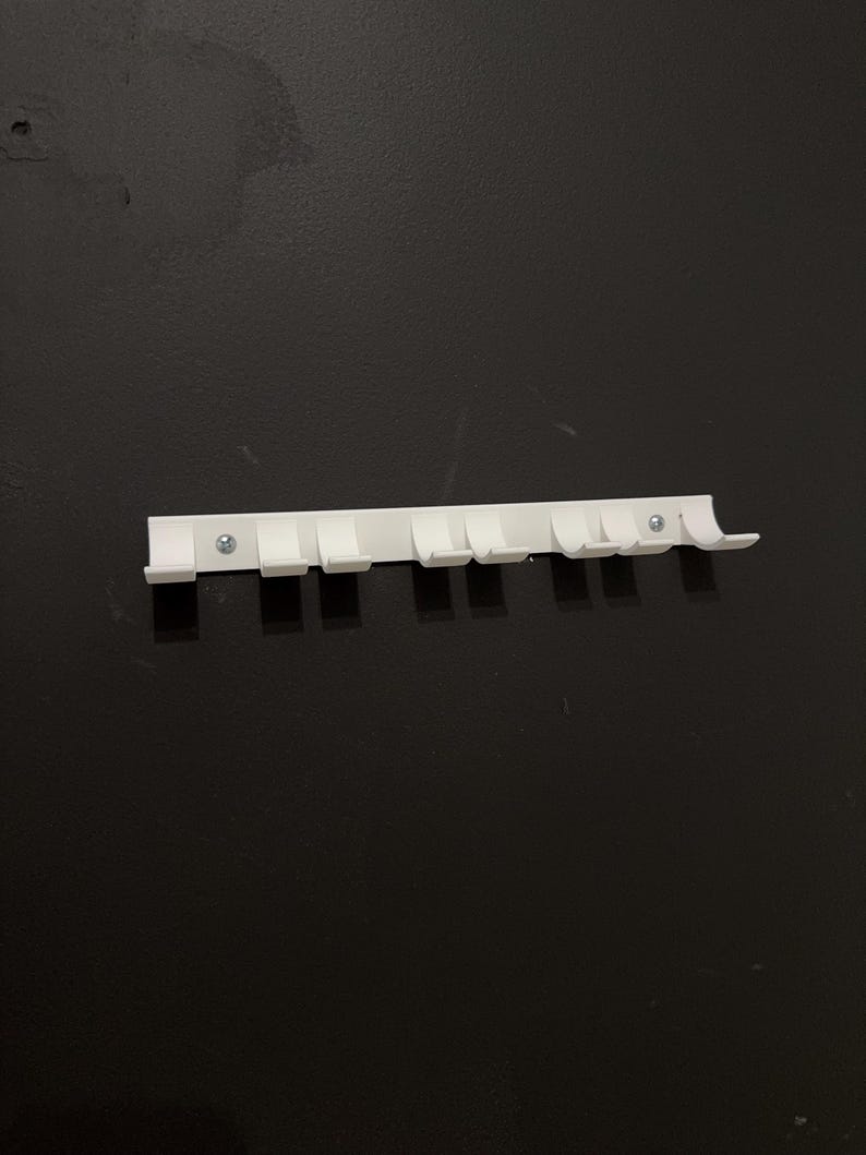 Puede incluir: Un organizador de pared de pl&aacute;stico blanco con siete ganchos. El organizador es rectangular y tiene una serie de ganchos peque&ntilde;os y espaciados para colgar objetos. Dos tornillos lo aseguran a la pared negra.