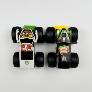May include: Two toy monster trucks, one white with a zombie face and the other black with "Grave Digger" and "Bad Bone" graphics. Both have large black tires and are set against a white background.