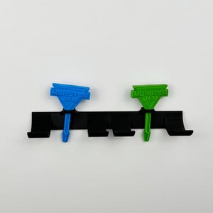 May include: A black plastic organizer with four slots, holding two Monster Jam-branded, colorful accessories. One is blue, the other green. The text "MONSTER JAM" is printed on each accessory. The background is plain white.