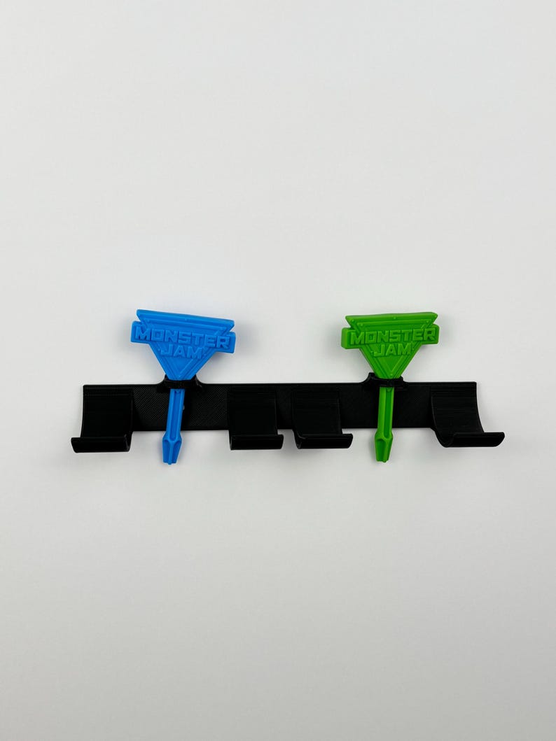 May include: A black plastic organizer with four slots, holding two Monster Jam-branded, colorful accessories. One is blue, the other green. The text "MONSTER JAM" is printed on each accessory. The background is plain white.