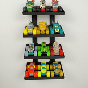 May include: A black wall-mounted shelf unit displaying a collection of toy monster trucks. The trucks are various colors, including red, green, yellow, and blue. Each truck has large, oversized tires. The shelf unit has four tiers.
