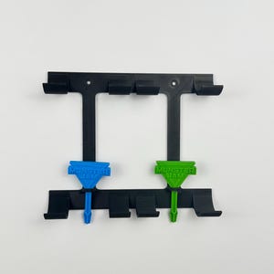 May include: Black plastic wall mount with multiple slots and two colored accents. The accents are blue and green, each with the text "MONSTER JAM". The mount is designed to hold items.