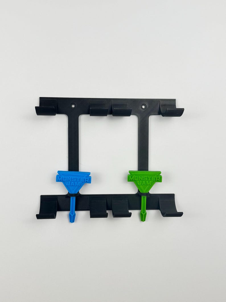 May include: Black plastic wall mount with multiple slots and two colored accents. The accents are blue and green, each with the text "MONSTER JAM". The mount is designed to hold items.