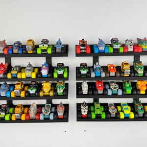 May include: Collection of colorful toy monster trucks displayed on black wall-mounted shelves. Each truck features unique designs and vibrant colors, including red, blue, green, and yellow. The collection is arranged on three shelves.