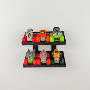 May include: A display of six toy monster trucks in various colors, including red, orange, green, and yellow. The trucks are arranged on two black shelves, each holding three trucks. The trucks have large tires and detailed designs.