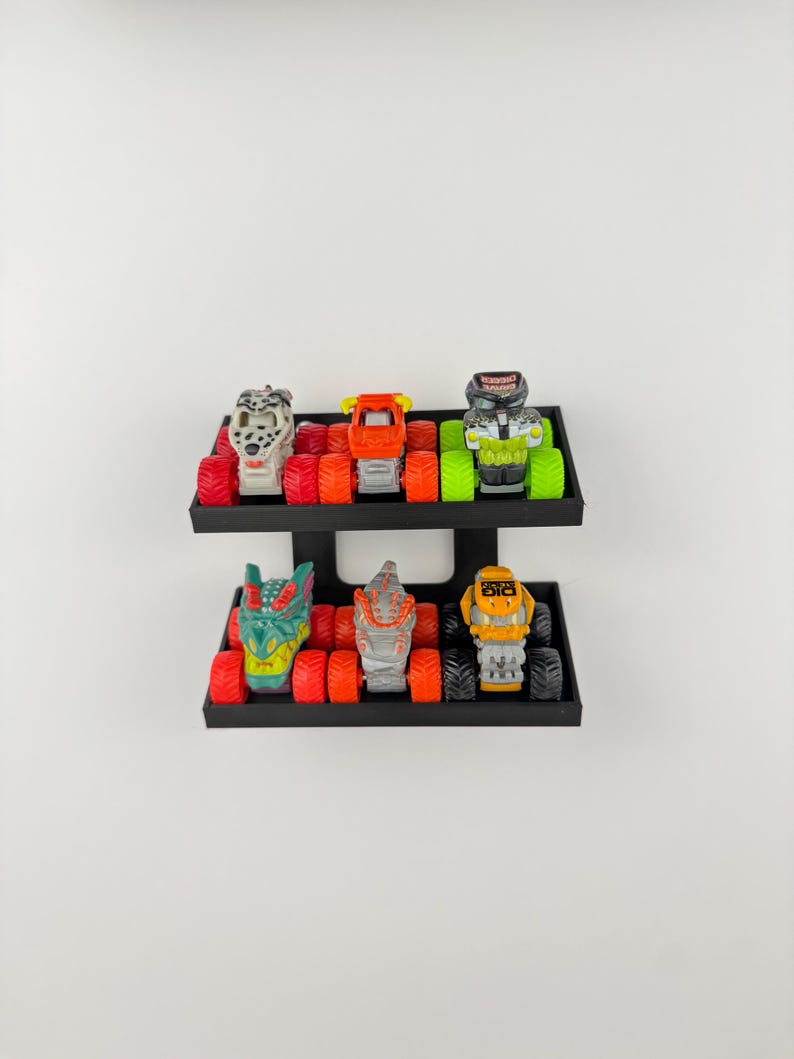 May include: A display of six toy monster trucks in various colors, including red, orange, green, and yellow. The trucks are arranged on two black shelves, each holding three trucks. The trucks have large tires and detailed designs.