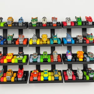 May include: A collection of colorful toy monster trucks displayed on black wall shelves. The trucks feature various designs and colors, including red, blue, green, and yellow. The shelves are mounted on a white wall.