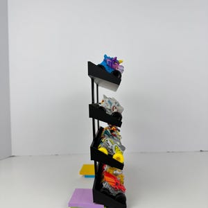 May include: A black display stand holding several toy monster trucks. The trucks are various colors, including blue, white, yellow, and red. The stand has three shelves and is set against a white background.