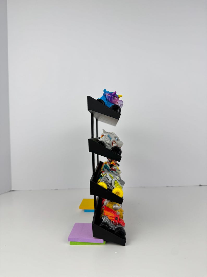 May include: A black display stand holding several toy monster trucks. The trucks are various colors, including blue, white, yellow, and red. The stand has three shelves and is set against a white background.