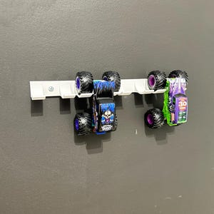 Puede incluir: Dos monster trucks de juguete, uno azul y otro verde, montados en un soporte blanco. El cami&oacute;n azul tiene un gr&aacute;fico de calavera y la palabra "Digger". El cami&oacute;n verde tiene el n&uacute;mero "30" y otros gr&aacute;ficos.