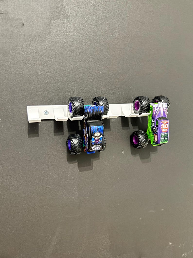 Puede incluir: Dos monster trucks de juguete, uno azul y otro verde, montados en un soporte blanco. El cami&oacute;n azul tiene un gr&aacute;fico de calavera y la palabra "Digger". El cami&oacute;n verde tiene el n&uacute;mero "30" y otros gr&aacute;ficos.