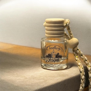 May include: A clear glass car air freshener bottle with a ribbed wooden cap and a braided beige cord featuring gold metallic threads. The label displays a circular logo with a candle illustration, pine branches, and the text Candles by Joseph Lyman, Maine.