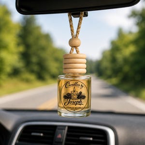 May include: A small, clear glass car air freshener bottle with a light wooden cap and a braided hanging cord. The bottle features a black circular label with a candle illustration, pine cones, and the text Candles by Joseph Lyman, Maine.