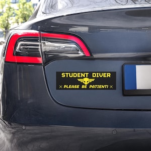 Könnte beinhalten: Ein schwarz-gelber Aufkleber auf einem grauen Auto mit der Aufschrift "STUDENT DIVER" über einer Totenkopf- und Flügelgrafik und dem Text "PLEASE BE PATIENT!" darunter.