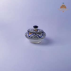 May include: A small, white ceramic jar with a lid. The jar and lid are decorated with blue, yellow, and black geometric patterns. The lid has a dark blue knob. The jar is likely used for storing small items or as a decorative piece.
