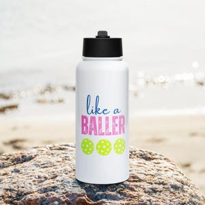 May include: A white water bottle with a black lid featuring the blue text like a and the pink text BALLER above three lime green pickleball graphics. The design is centered on the front of the cylindrical container, which has a smooth finish.