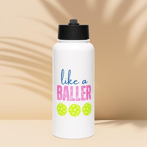 May include: A white water bottle with a black lid featuring the blue text like and the pink distressed text BALLER above three lime green pickleball graphics. The design is centered on the front of the cylindrical container.