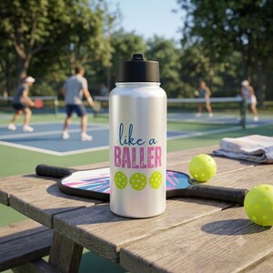 May include: A silver water bottle with a black lid featuring the blue text like a and pink text BALLER above three lime green pickleball icons. The bottle sits on a wooden surface next to a pickleball paddle and two bright yellow pickleballs.