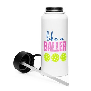 May include: A white water bottle with a black lid and a clear straw. The bottle features the blue text "like a" above the pink distressed text "BALLER" and three lime green pickleball icons arranged in a horizontal row on the front surface.