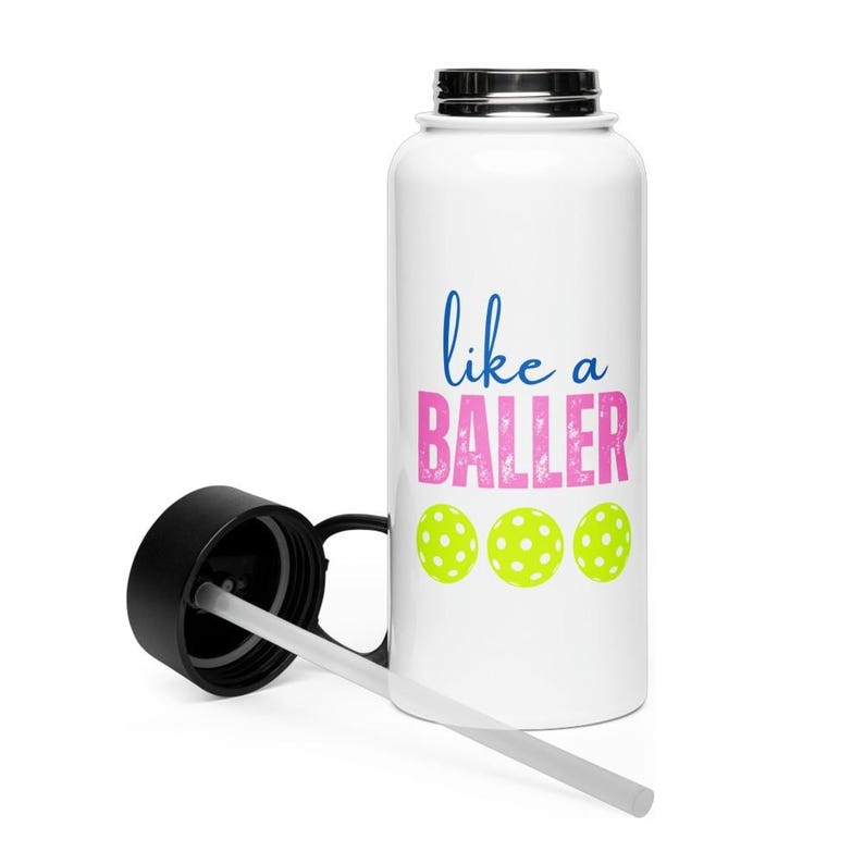 May include: A white water bottle with a black lid and a clear straw. The bottle features the blue text "like a" above the pink distressed text "BALLER" and three lime green pickleball icons arranged in a horizontal row on the front surface.