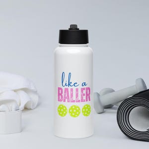 May include: A white water bottle with a black screw-top lid. The front features the blue text "like a" above the pink text "BALLER" in a distressed font, followed by three bright yellow pickleballs with white circular patterns.