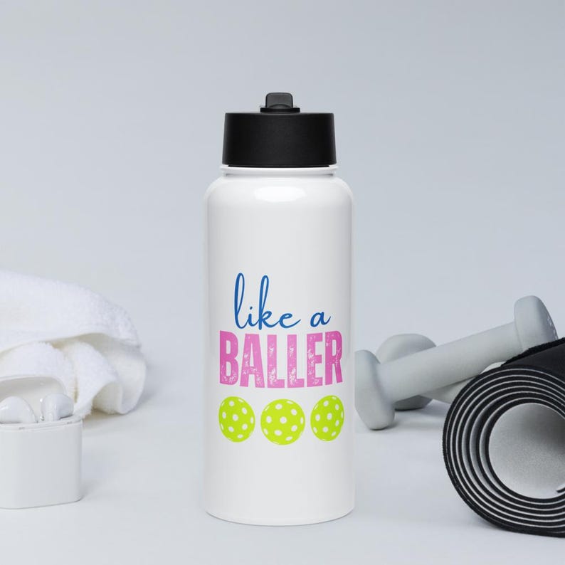 May include: A white water bottle with a black screw-top lid. The front features the blue text "like a" above the pink text "BALLER" in a distressed font, followed by three bright yellow pickleballs with white circular patterns.