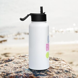 May include: A white cylindrical water bottle with a black lid and a flip-up straw. The bottle features text in blue and pink, along with a small lime green circular graphic. The design is printed vertically on the side of the white container.