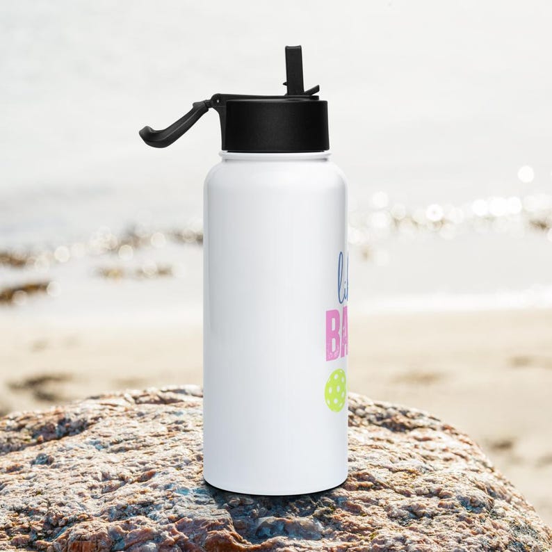 May include: A white cylindrical water bottle with a black lid and a flip-up straw. The bottle features text in blue and pink, along with a small lime green circular graphic. The design is printed vertically on the side of the white container.