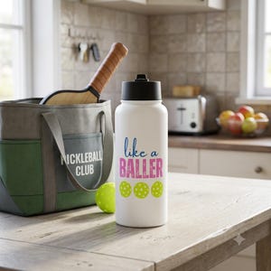 May include: A white water bottle with a black lid featuring the blue text like a and pink text BALLER above three lime green pickleballs. Next to it is a grey and green tote bag with the text PICKLEBALL CLUB and a pickleball paddle handle.