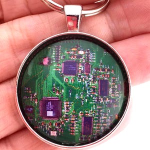 May include: A circular keychain featuring a silver-toned metal frame and a domed glass cabochon. The insert displays a detailed, high-resolution graphic of a green computer circuit board with various purple integrated chips and electronic components.