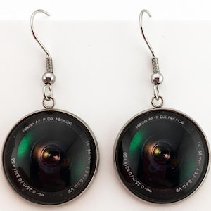 May include: A pair of silver-toned hook earrings featuring circular glass cabochons. Each cabochon displays a detailed graphic of a camera lens with the text Nikon AF-P DX NIKKOR 18-55mm 1:3.5-5.6G VR and 0.25m/0.82ft printed around the dark lens element.